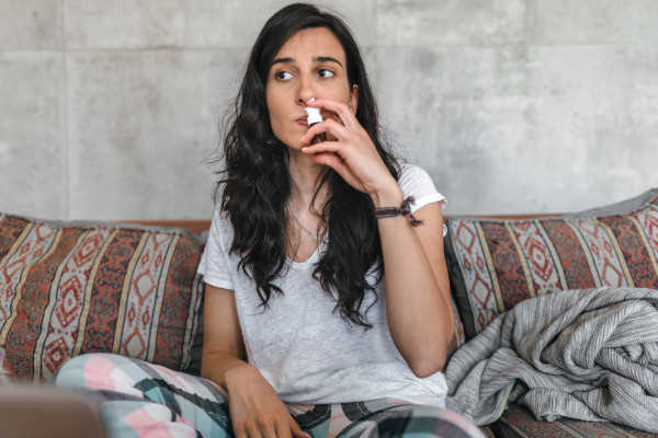 A woman sitting on the cough with a blanket, using a nasal spray for nasal congestion
